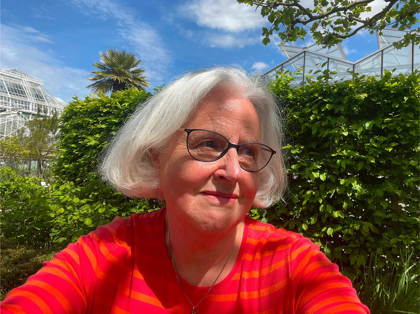 Ilka Scholz mit grauem Bob und Brille in einem sonnigen Garten, im Hintergrund Gewächshäuser.